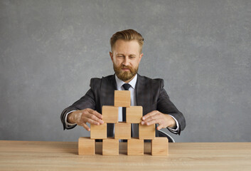 Man in suit building pyramid on office table. Ambitious male business leader sitting at desk and...