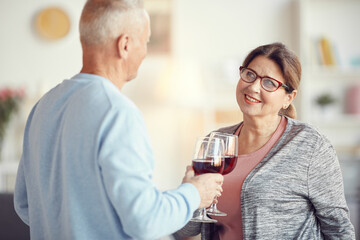 Positive senior couple in casual clothing standing in living room and toasting with wineglasses while celebrating anniversary