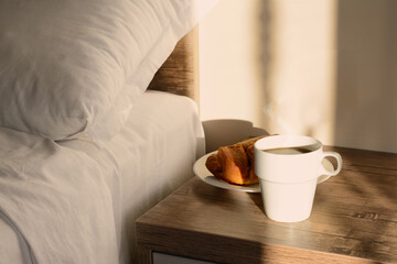 Fototapeta na wymiar Cup of coffee and croissant on night stand near bed in morning