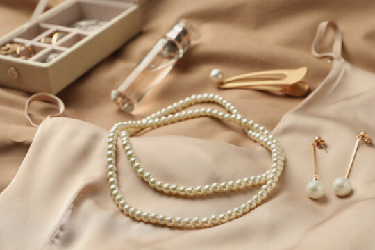 Stylish jewelry with pearls, luxury perfume and silk dress on beige fabric, closeup