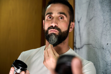 Young man applying wax and oil to his beard in front of mirror