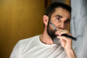 Young man with a beard shaving with a traditional knife in front of the mirror