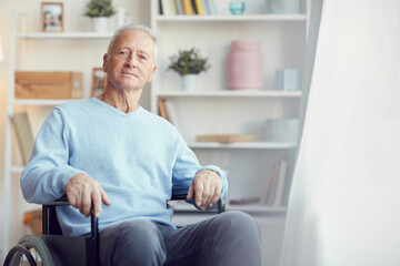 Portrait of content handicapped senior man in light blue sweater using wheelchair for motion at home