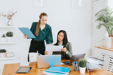 two young business women professionals in formal wear clothes work in modern office using laptop, tablet, brainstorm and search for solutions together, confident independent Asian girl solves problems