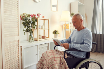 Serious senior man in sweater sitting with covered legs in wheelchair and using tablet while searching for information about own injury