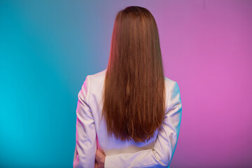 Woman with long hair standing back portrait with neon lights colors effect. Female model isolated on neon colored background wearing white suit.