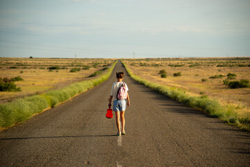 Obraz premium A girl walks along an empty road in the steppe. She has a ukulele in her hands and a backpack on her shoulders. It's summer and warm outside. Open road.