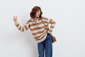 a cheerful woman standing on a white background in a striped sweater put her hand on the belt, turning her head to the side, raising her other hand up and slightly bent it