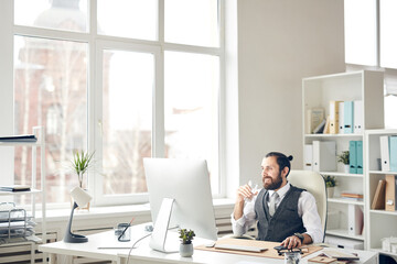 Cheerful confident young hipster businessman with ponytail sitting at table and drinking water while working with computer