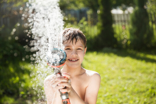 Happy Kid Playing With Garden Hose And Having Fun With Spray Of Water In Sunny Backyard. Summer Time. Kid Boy Helps Water Garden With Hose. Slow Life. Enjoying The Little Things. Summer Holiday