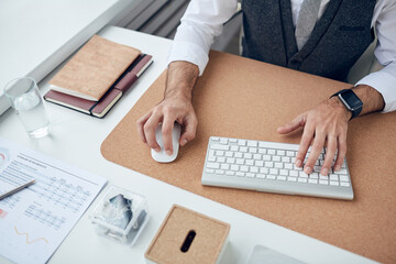 Close-up of unrecognizable businessman with smart watch sitting at desk and using wireless mouse and keyboard