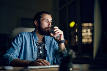 Serious thoughtful young bearded man in casual shirt sitting in night office and drinking water while using computer