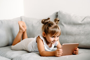 Toddler girl playing with digital wireless tablet computer on couch at home. Baby child growing with online applications. Child and electronic devices concept. Portrait of toddler with smartphone. 