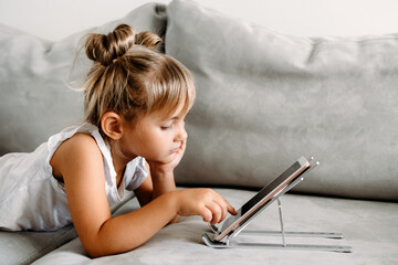 Toddler girl  lies on couch at home and playing with digital wireless tablet computer. Child and electronic devices concept. Portrait of toddler with smartphone. Education and learning concept.