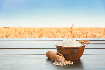 Bowl of wheat flour and scoop with grains on wooden table in field