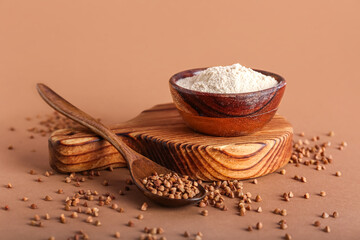 Board with bowl of flour, spoon and buckwheat grains on brown background