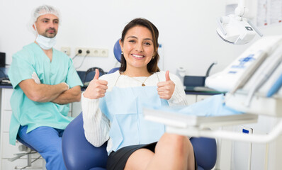 Happy cute latin woman sitting in dental chair after teeth cure giving thumb up