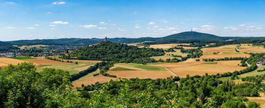 Panoramafoto vom Bergfried der H&ouml;henburg Vetzberg auf einem Vulkanberg in Mittelhessen, eingebettet in eine landwirtschaftlich genutzte Landschaft mit leicht bew&ouml;lktem Himmel am Horizont und dem D&uuml;nsb