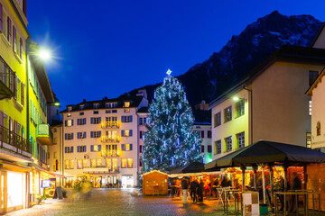 Night view of pedestrian cobbled street of Brig town with traditional festive fair and Christmas tree decorated with bright lighting on background of snow covered Alpine peaks, Valais, Switzerland.