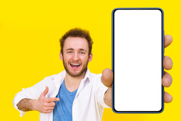 Check this app young man pointing at smartphone showing a white empty screen game, bet, lottery win isolated over yellow background. Product placement for mobile application advertisement