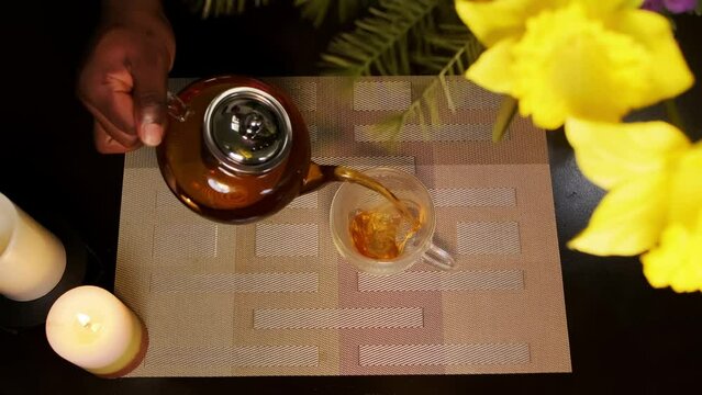 Top down shot of an African American man pouring steaming hot tea into a heart shaped transparent glass tea cup sitting on a cozy dining table.