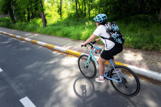 Young girl in helmet with backpack rides bicycle on asphalt road at summer day.