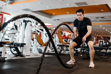 Athletic Asian young man doing some fitness exercises with battle rope at fitness gym. Muscular powerful aggressive training, Concept for exercising, fitness, and healthy lifestyle.