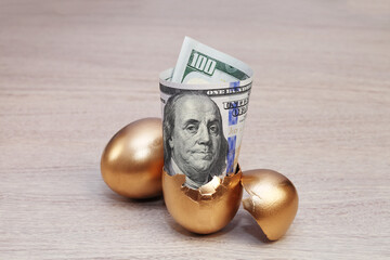 Dollar bills in golden broken egg on wooden background.