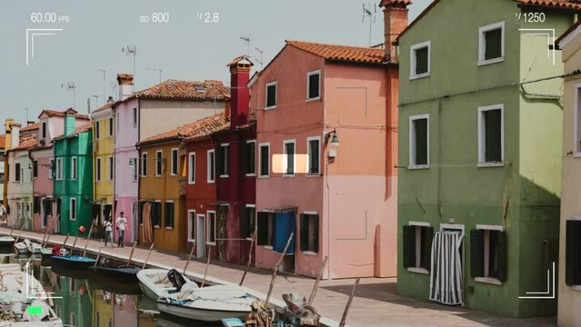 Burano, Venice, Italy - July 2, 2022: A look through the photo camera at brightly colored houses and water canal with boats in Burano, people walk and rest on streets