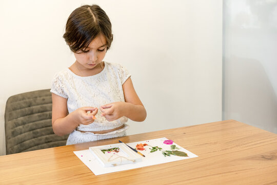 Child with white paper, leaves, and flowers making herbarium on dining wood table in home. Dried flower art.