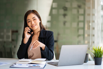 Successful young Asian businesswoman holding a smartphone working on a laptop computer financial graph data on the desk in the office.