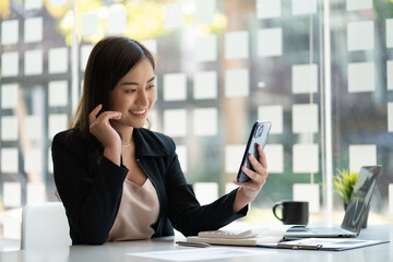 Successful young Asian businesswoman holding a smartphone working on a laptop computer financial graph data on the desk in the office.