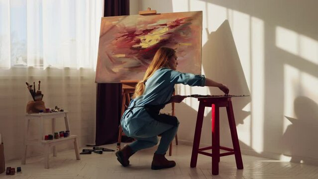 Young woman artist painting in art studio. Female artist is working at picture using oil paints and palette-knife creating beautiful paints a picture. Painting technique and painters concept.