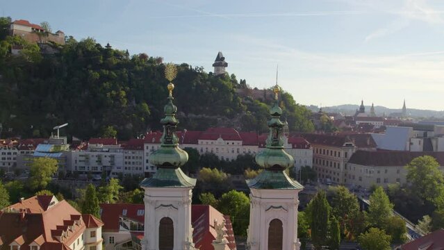 Aerial view flying between Minoritenkirche baroque towers towards Uhrturm hilltop clock tower Graz's Schlo&szlig;berg