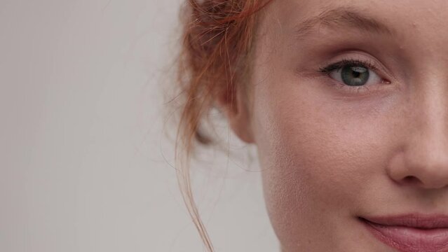 Close up view of beautiful young woman touching her half face and look at the camera. Portrait pretty ginger female with nude make up posing at studio background. Concept of cosmetology, skincare.