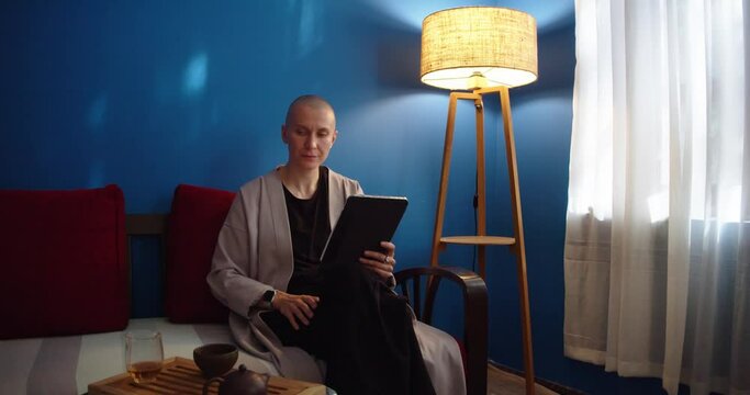 Stylish woman sitting on sofa using ipad and drinking tea at home in evening. Pretty female scrolling tablet screen and reading by lamplight. Shopping online