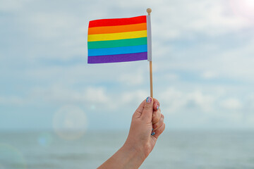 Rainbow Flag. Woman hand holding the flag rainbow colors. LGBT Pride. Rainbow flag symbol of bisexual people, gays and lesbians LGBT, LGBTQ. Good for Party. Sunny Day. Ocean wave on background.