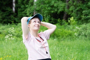 Young woman in pink shirt and jeans cap, headphones stay in forest glade and dreams with two hands...