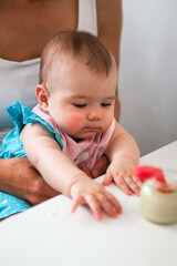 Naklejka premium Child six month old with mother on a kitchen at a table with jar with vegetable puree and spoon. Baby girl tries to eat herself.