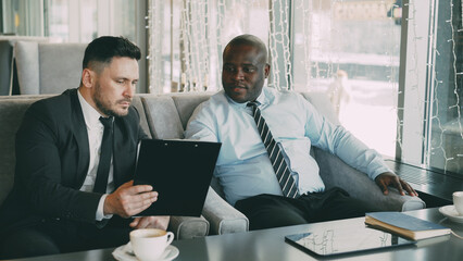 Two multi-ethnic businessmen looking at clipboard and discussing startup projects in modern cafe during lunch time
