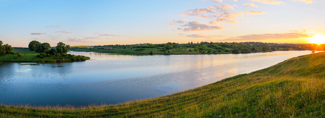 Summer panoramic rural landscape with green hills and blue calm river.Beautiful clouds in colorful sunrise sky over the land.