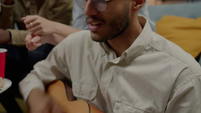 Close-up of creative mixed race man playing the guitar and singing while friends listening at home. Musical instrument and youth culture concept.