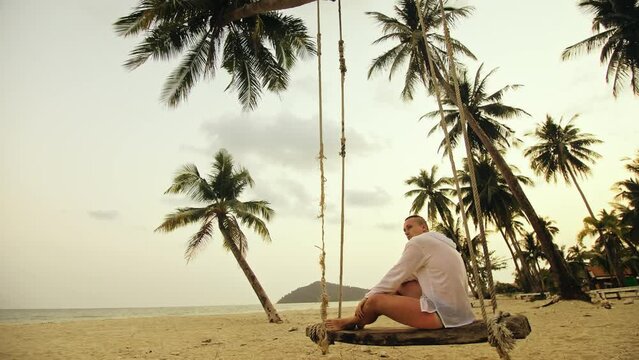 Cute loving couple on the swing on tropical beach. Woman and man in white shirt enjoy gold sunset and life in beaches, palm tree. Evening warm sunset. Summer holiday vacation tropical tourism concept