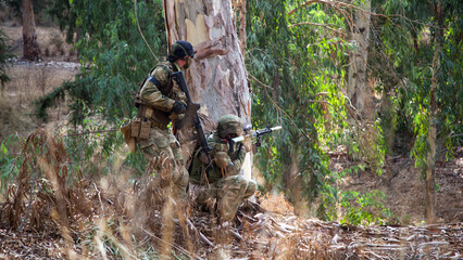 Two airsoft players hid behind a tree and fire at the enemy covering each other. Two guys with weapons in camouflage are hiding behind a tree.