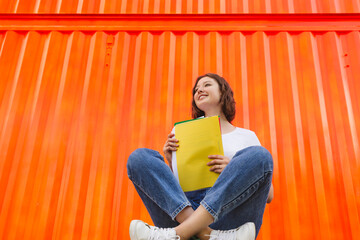 Smiling teenage girl with yellow file folder sitting cross-legged in front of orange cargo container