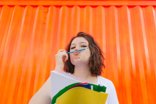Teenage girl with blue pencil puckering lips in front of orange cargo container