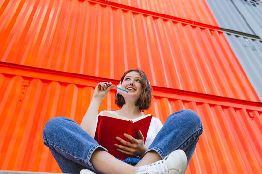 Smiling thoughtful teenage girl with red notebook sitting in front of orange cargo container