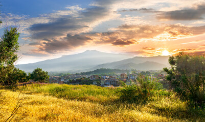 view from hill with golden grass and green bushes to a valley town with majectic mountains and scenic cloudy sunset on background