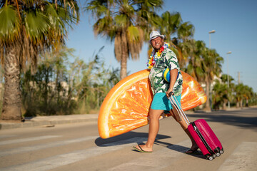 Smiling man holding orange slice shaped pool raft and suitcase crossing road