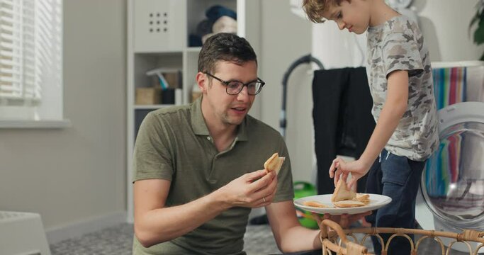 Man and son sit on laundry room floor relaxing after sorting laundry, eating cheese toast snack during lunch.